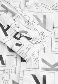 Balta krekla ar pogām, kurā ir drukāta melnu ģeometrisku burtu un formu raksts. Pogas uz aprocēm ar teksturētu audumu.