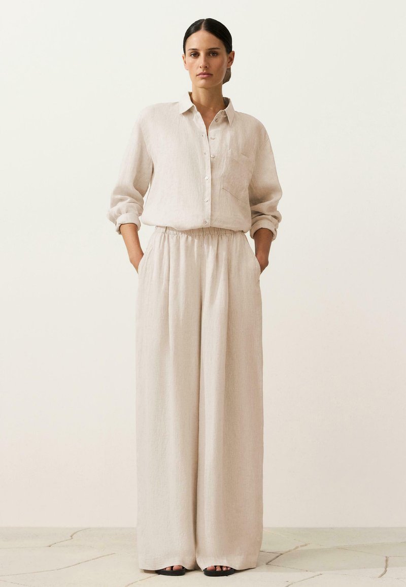 Femme portant une chemise ample beige à manches longues boutonnée, rentrée dans un pantalon ample assorti, mains dans les poches, debout sur un sol carrelé.