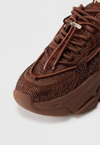 Brun sneaker med en tekstureret overdel med indlejrede glimmer, rund tå og detaljeret snøresystem. Logo detaljeret på siden.