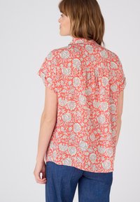 Camisa de manga corta color coral con un patrón floral en blanco, un corte holgado y un dobladillo curvado. La tela parece ligera y transpirable.