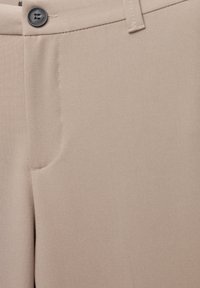 Détail d’un pantalon beige montrant le bouton, la ceinture, la passants de ceinture et la couture de la poche avant sur un tissu lisse.