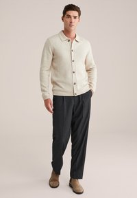 Cardigan din lână bej, cu guler și cinci butoane mari maro, asortat cu pantaloni gri închis, croiți, și ghete din suede maro, cu glezna desfăcută.