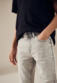 Jean en denim gris clair avec une coupe ajustée, un léger effet froissé, et une fermeture à bouton, associé à une chemise noire à manches courtes.