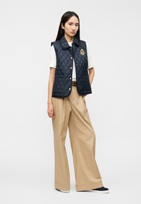 Σκούρο μπλε γιλέκο με καπιτονέ σχέδιο και γιακά, διακοσμημένο με λογότυπο στο στήθος, φορεμένο πάνω από λευκό πουκάμισο και χακί παντελόνι με φαρδιά μπατζάκια.