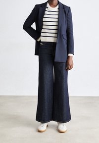 Blazer bleu marine sur un pull blanc à rayures, associé à un jean denim large foncé et des baskets blanches à semelle marron clair.