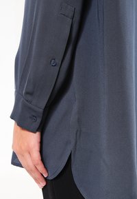 Chemise bleu marine à boutons, en tissu texturé, avec des manches longues et un ourlet arrondi. Les boutons se trouvent sur les poignets.