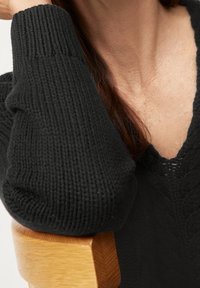 Svart stickad tröja med ribbade ärmslut, med en djup V-ringning och texturerat mönster. En trästolsarm synlig i förgrunden.