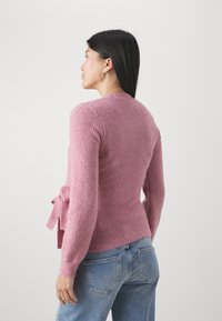 Rosa ribbad cardigan med långa ärmar, en knytning vid midjan och en åtsittande silhuett, tillsammans med ljusblå jeans.