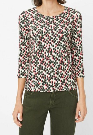 Vrouw draagt een wit topje met zwarte en roze abstracte vierkante patronen en driekwart mouwen, gecombineerd met donkergroene broek.