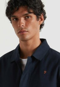 Camicia colletto blu navy in cotone, con un piccolo logo ricamato arancione. Tessuto liscio, vestibilità standard.