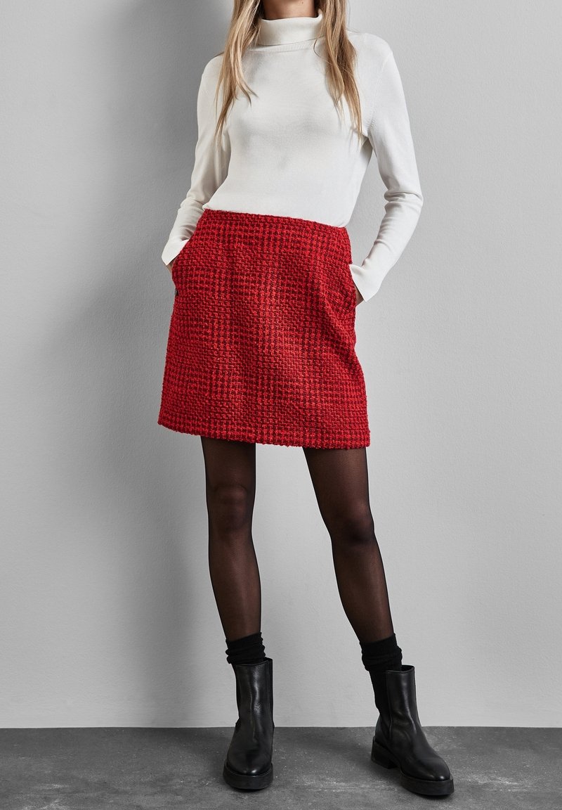 Röd texturerad kjol med fickor, bärs över svarta strumpbyxor och svarta ankelskor, matchat med en vit långärmad topp.