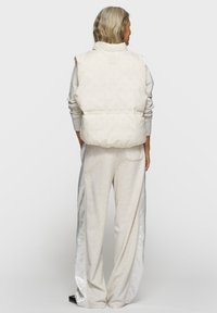 Steppweste in off-weißem Steppstoff mit hohem Kragen und elastischem Bund, kombiniert mit hellgrauen Jogginghosen mit Satin-Seitenstreifen.