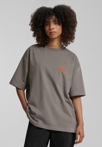 Camiseta gris de gran tamaño hecha de algodón, con mangas cortas, cuello redondo y un logo bordado en naranja en el lado izquierdo del pecho.