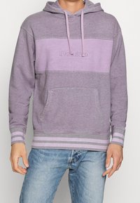 Sweat à capuche violet avec un bloc de couleur central, un logo brodé et une poche avant ; présente des poignets et un ourlet rayés en gris clair.