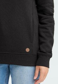 Sweatshirt noir avec des poignets côtelés, une poche avant et un petit patch logo en cuir marron. Tissu doux au toucher lisse.