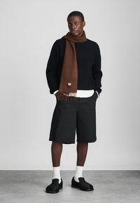 Maglione nero a coste, sciarpa marrone con frange, pantaloni corti neri e scarpe nere imbottite. La modella si trova su uno sfondo neutro.