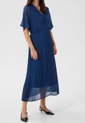 Nainen, joka pitää polvipituista laivastonsinistä pliseerattua mekkoa, jossa on lyhyet läpikuultavat hihat, sekä mustat matalakorkoiset slingback-kengät, seisoo harmaalla lattialla.