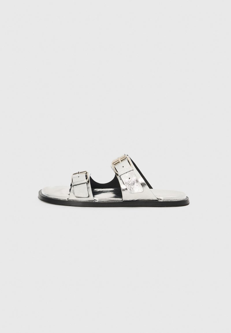 Sølvmetallic slide sandaler med to justerbare spænder, sort indersål og flad gummiydersål. Glat tekstur og åben design.