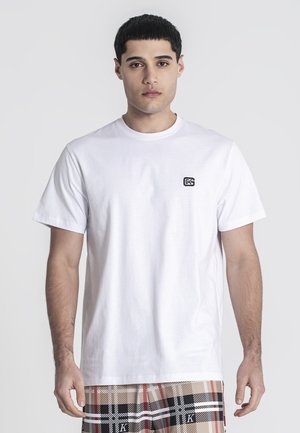 Camiseta blanca de algodón con cuello redondo, mangas cortas y detalle de logo en el lado izquierdo del pecho. Combinada con pantalones cortos de cuadros con patrones en negro, rojo y beige.