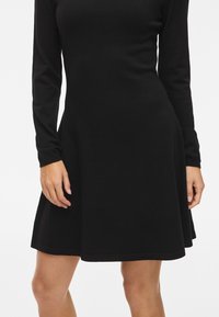 Rochie neagră cu mâneci lungi, cu un corset ajustat și fustă evazată. Confectionată dintr-un material moale, având un decolteu rotund și o margine simplă.