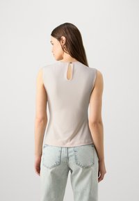 Top sin mangas de color beige claro con un detalle de ojal redondo y cierre de botón en la parte posterior. Tela suave y elástica con una forma ajustada.