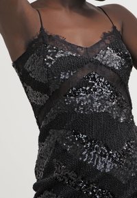 Gros plan sur une personne portant une robe noire à fines bretelles, décorée de sequins noirs et argentés ainsi que de motifs en dentelle.