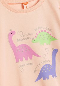 Różowa bawełniana koszula z trzema kolorowymi grafikami dinozaurów w odcieniach fioletu, zieleni i różu, z brokatowymi akcentami i zabawnym napisem.