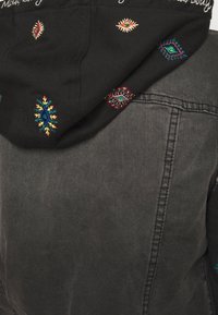 Svart huvtröja med färgglada broderade mönster, inklusive geometriska former och ögonmotiv, i lager över en mörk jeansjacka.