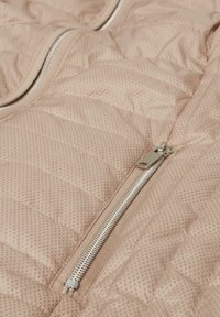 Beige gesteppte Weste mit einem mittig platzierten silbernen Reißverschluss und einem diagonalen Muster. Glattes Material mit abgerundeten Kanten.