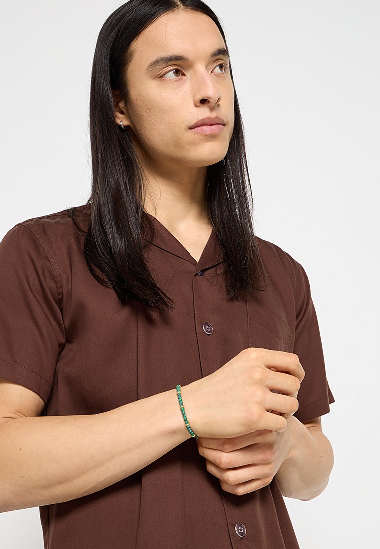 Chemise brune à manches courtes avec col et boutons, associée à un bracelet en perles aux teintes turquoise et verte porté au poignet.