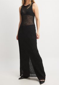 Donna che indossa un maxi dress black senza maniche in crochet con motivi a diamante e spacco laterale, abbinato a scarpe nere con punta.
