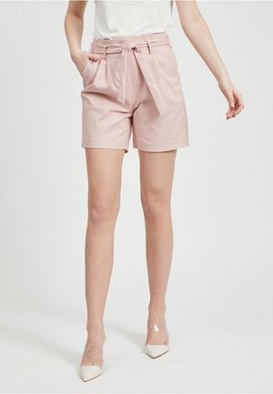 Frau in hellrosa Hochbund-Shorts mit gebundenem Gürtel, weißem Oberteil und weißen, spitzen Absätzen mit transparenten Riemen, stehend vor weißem Hintergrund.