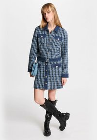 Vestido de mezclilla con un patrón de pata de gallo azul, que incluye bolsillos, botones plateados, cintura ajustada y un cuello. Botas negras hasta la rodilla.