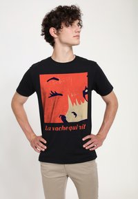 Sort bomulds T-shirt med et farverigt grafisk print af et kogehoved og teksten "la vache qui rit" i rød. Korte ærmer, rund halsudskæring.