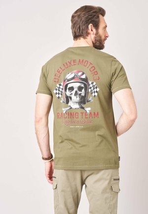Man draagt een olijfgroen T-shirt met een schedel in een vintage racehelm en geruite vlaggen, tekst "Deluxe Motors Racing Team."