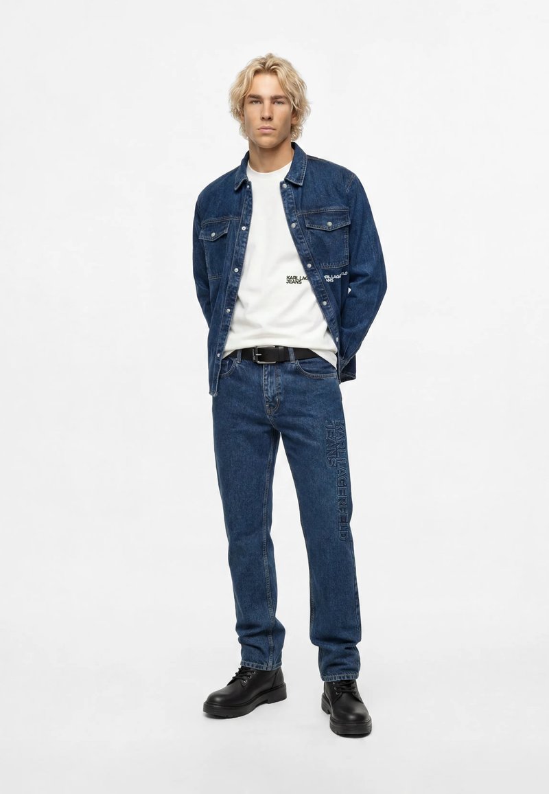 Jeune homme aux cheveux blonds portant une veste en jean bleu foncé et un jean, un t-shirt blanc, une ceinture noire et des chaussures noires à lacets, debout sur un fond blanc.