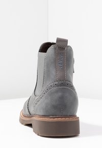 Šedá semišová kotníková bota s elastickými bočními panely, zaoblenou špičkou, dekorativním prošíváním a hnědou gumovou podrážkou. Název značky je na zadním poutku.