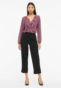 Oprijeta vijoličasta bluza z globokim V-izrezom in dolgimi rokavi, kombinirana z visokim pasom črnih hlač z zavihanimi robovi ter črnimi koničastimi petami.