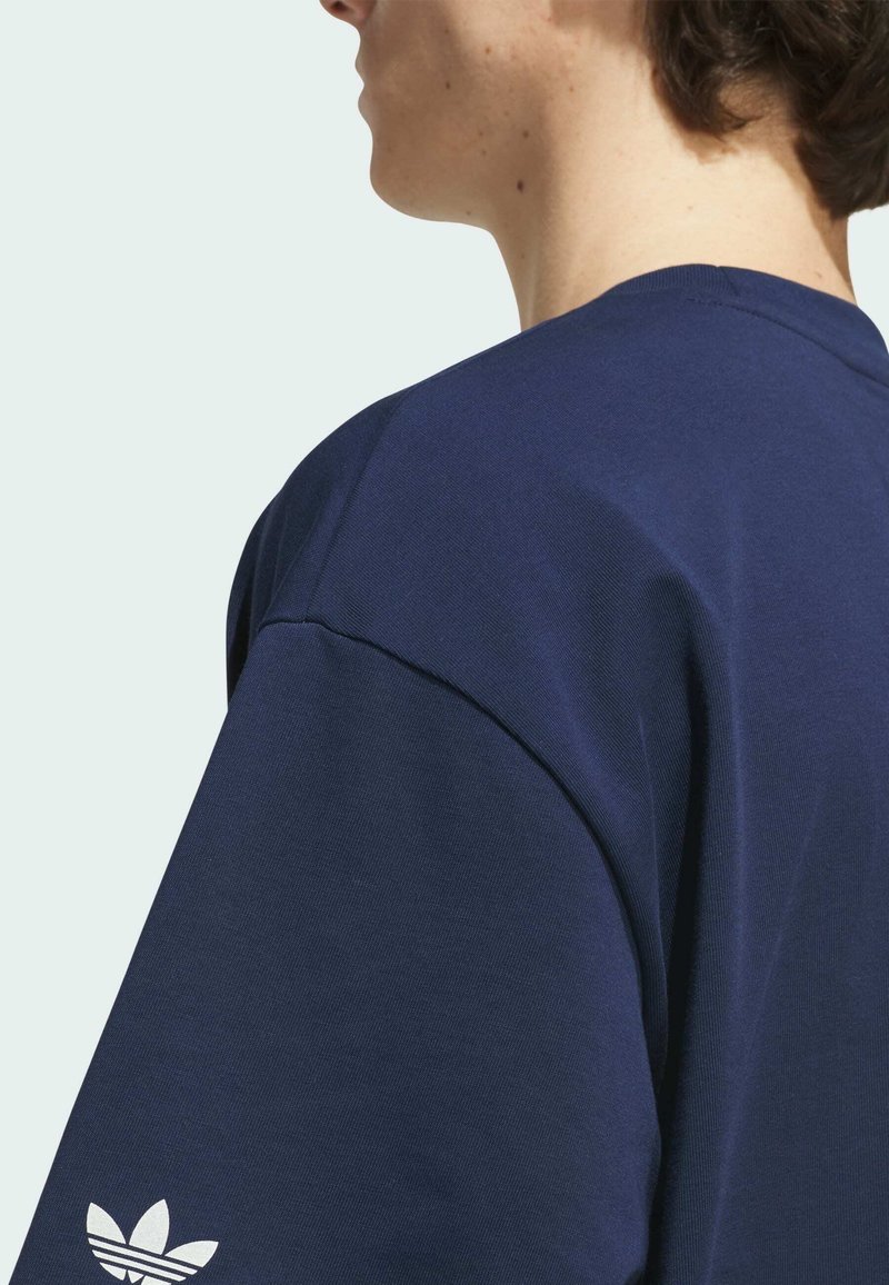 Personne portant une chemise bleu marine avec un logo Adidas blanc sur la manche, vue de côté et de dos, sur un fond clair.