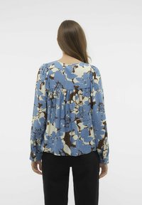 Helles blaues Bluse mit floralen Mustern und langen Ärmeln, mit braunen Akzenten und Raffung im Rücken. Weicher, strukturierter Stoff.