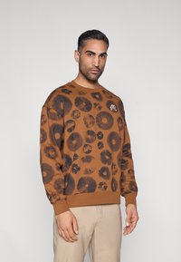 Καφέ φούτερ με μαύρο λουλουδένιο σχέδιο, ριμπ λαιμόκοψη και μανσέτες. Ο σχεδιασμός περιλαμβάνει λουλούδια διαφόρων μεγεθών.