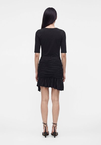 Femme aux longs cheveux raides portant un haut noir moulant, une mini-jupe noire froncée à ourlet à volants et des sandales à talons noires à brides, debout de dos.