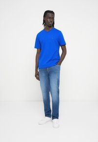 Μπλε βαμβακερό t-shirt με λαιμόκοψη σε σχήμα V, κοντά μανίκια, λεπτομέρεια με λογότυπο μπροστά. Συνδυασμένο με ανοιχτό μπλε τζιν και λευκά αθλητικά παπούτσια. Μίνιμαλ σχέδιο, χαλαρή εφαρμογή.