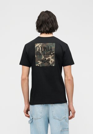 Άτομο με σκούρα μαλλιά μέχρι τους ώμους φοράει μαύρο T-shirt με παραμάνα παραλλαγής που γράφει "Alpha Industries" στην πλάτη και ανοιχτό μπλε τζιν.