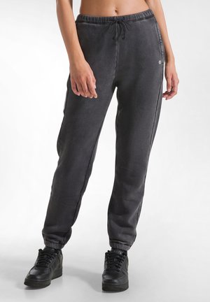 Schwarze Jogginghose mit verstellbarem Bund, schmal zulaufenden Säumen und glatter Textur, kombiniert mit schwarzen Sneakers. Logo-Detail auf der Hose.