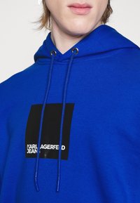Sudadera azul con un logo cuadrado negro que presenta texto blanco. Incluye cordones y un tejido texturizado. Vista en primer plano del escote.