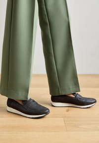 Svarta slip-on skor i textilerad material med vit sula, tillsammans med gröna vida byxor på en trägolv.