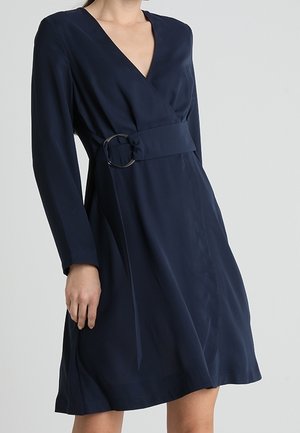 Vrouw draagt een marineblauwe wikkeljurk met lange mouwen en een brede riem met een grote cirkelvormige gesp in de taille.