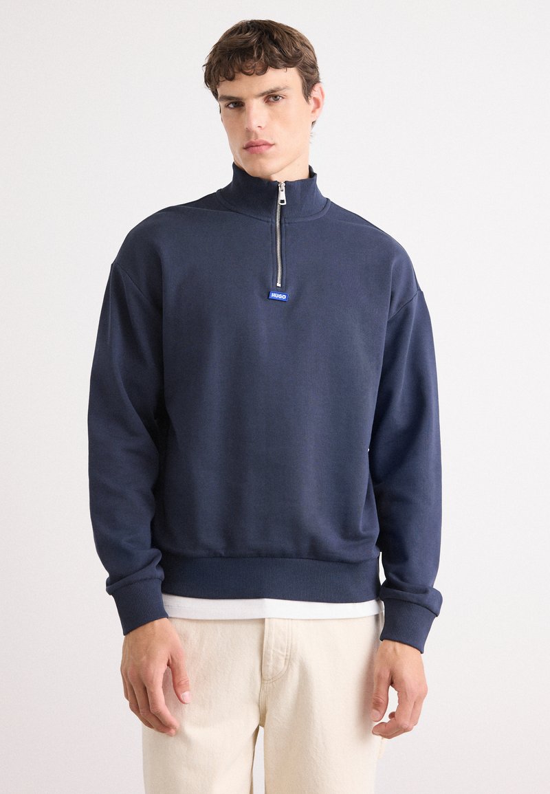 Sudadera con cremallera azul marino hecha de tela suave, con cuello alto, puños de canalé y un pequeño logotipo en el cuello.
