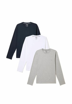 Mann trägt ein hellgraues langärmliges Shirt, blaue Jeans und olivgrüne Sneakers, steht vor einer schlichten hellen Wand.